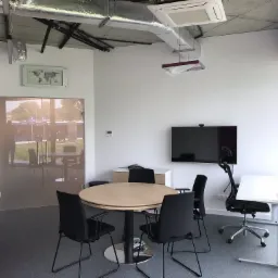 Jasne, nowocześnie urządzone biuro z okrągłym stołem konferencyjnym, krzesłami, biurkiem regulowanym, telewizorem na ścianie i mapą świata w ramce.