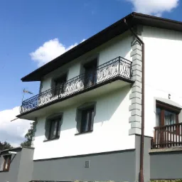 Dwukondygnacyjny dom z szarą podmurówką i białymi ścianami, z balkonami z kutą balustradą, widok z dołu na tle błękitnego nieba z chmurami.