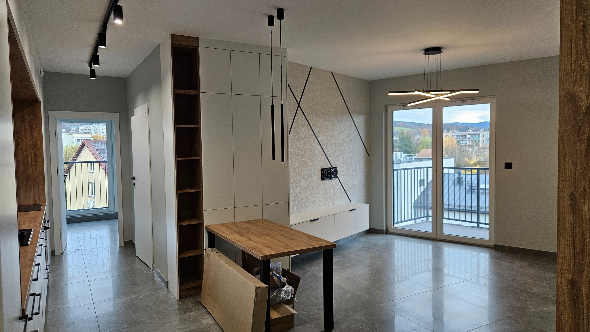 Nowoczesne wnętrze po remoncie: szare płytki podłogowe, białe meble, dekoracyjna ściana z geometrycznym wzorem, oświetlenie punktowe i wiszące lampy. Widok na balkon.