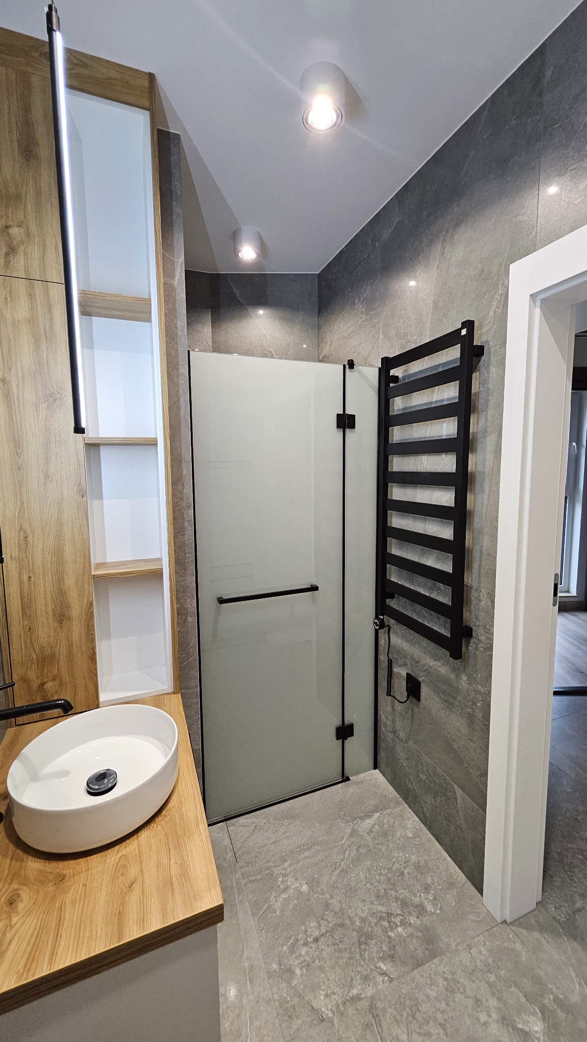 Nowoczesna łazienka z kabiną prysznicową z matowego szkła, grzejnikiem drabinkowym i umywalką nablatową na drewnianym blacie. Szare płytki imitujące kamień na ścianach i podłodze.