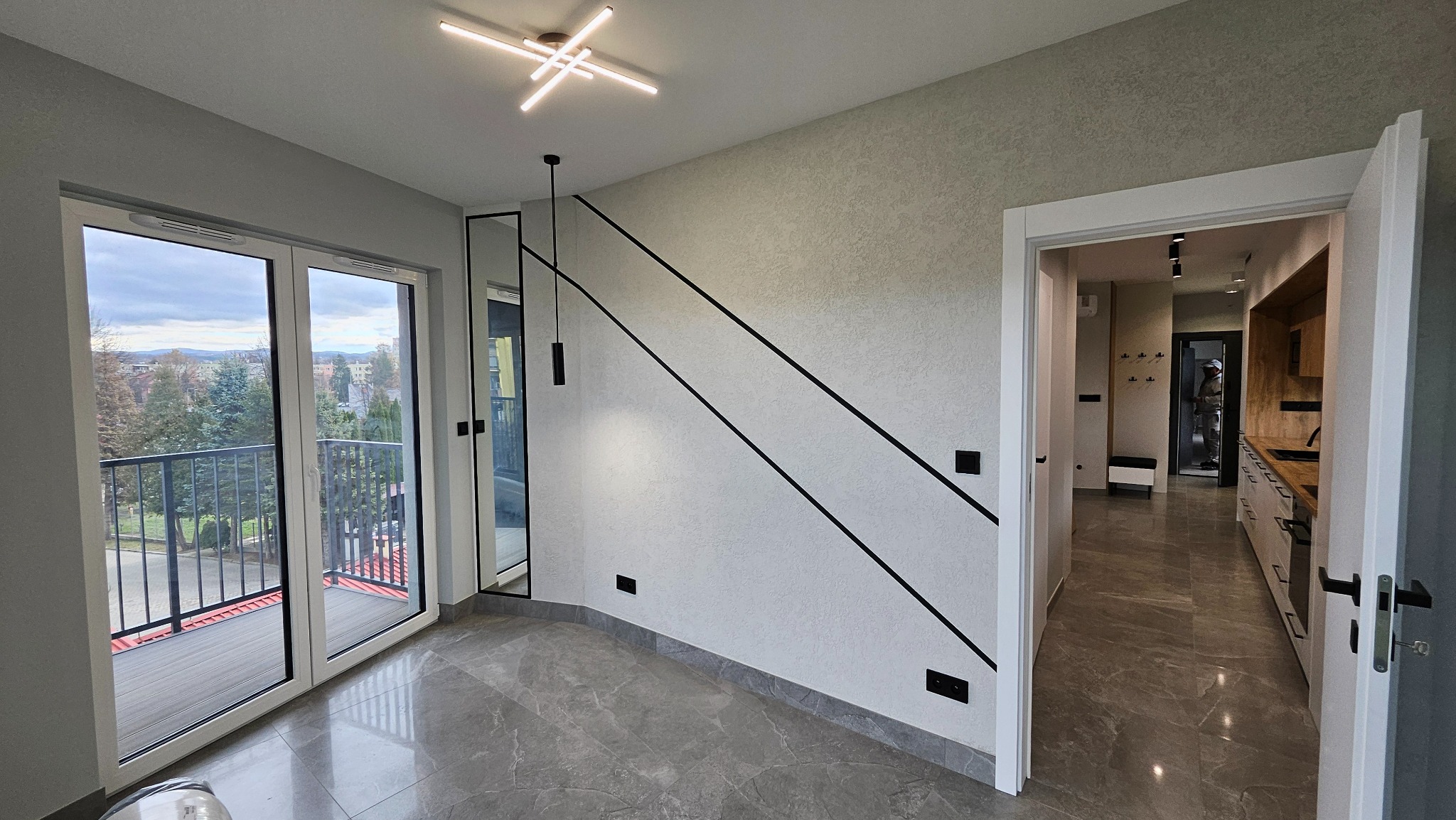 Nowoczesne wnętrze po remoncie: jasne ściany z dekoracyjnymi czarnymi pasami, szara podłoga, wyjście na balkon i widok na kuchnię. Minimalistyczny design.