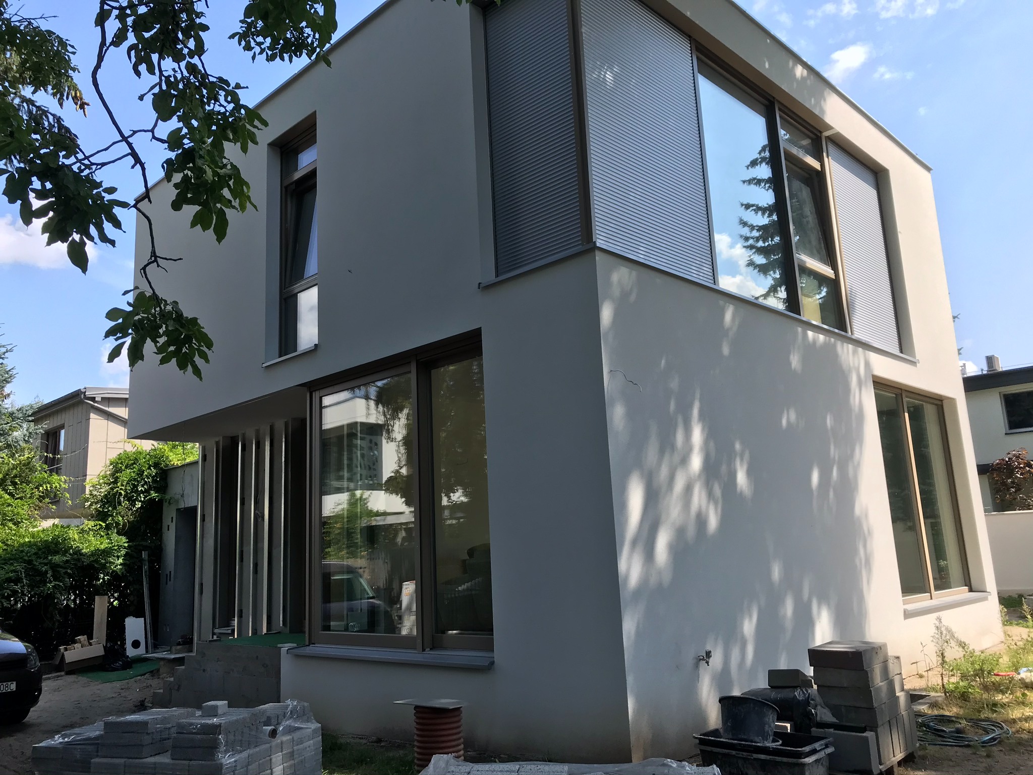 Nowoczesny dom z jasną elewacją, widoczne okna z roletami zewnętrznymi, teren wokół w trakcie prac wykończeniowych, paleta z kostką brukową na pierwszym planie.