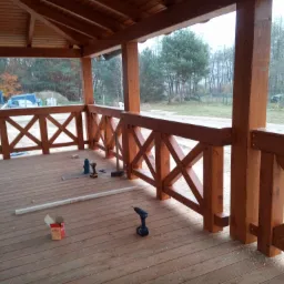 Drewniana altana w trakcie budowy w Bytowie, widoczne narzędzia: wiertarki, skrzynka po wkrętach, drewniana podłoga i balustrada, w tle las.