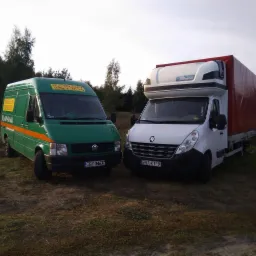 Dwa busy: zielony VW LT z pomarańczowym pasem i napisem reklamowym oraz biały Renault Master z plandeką, stojące na trawiastym terenie pod lasem.
