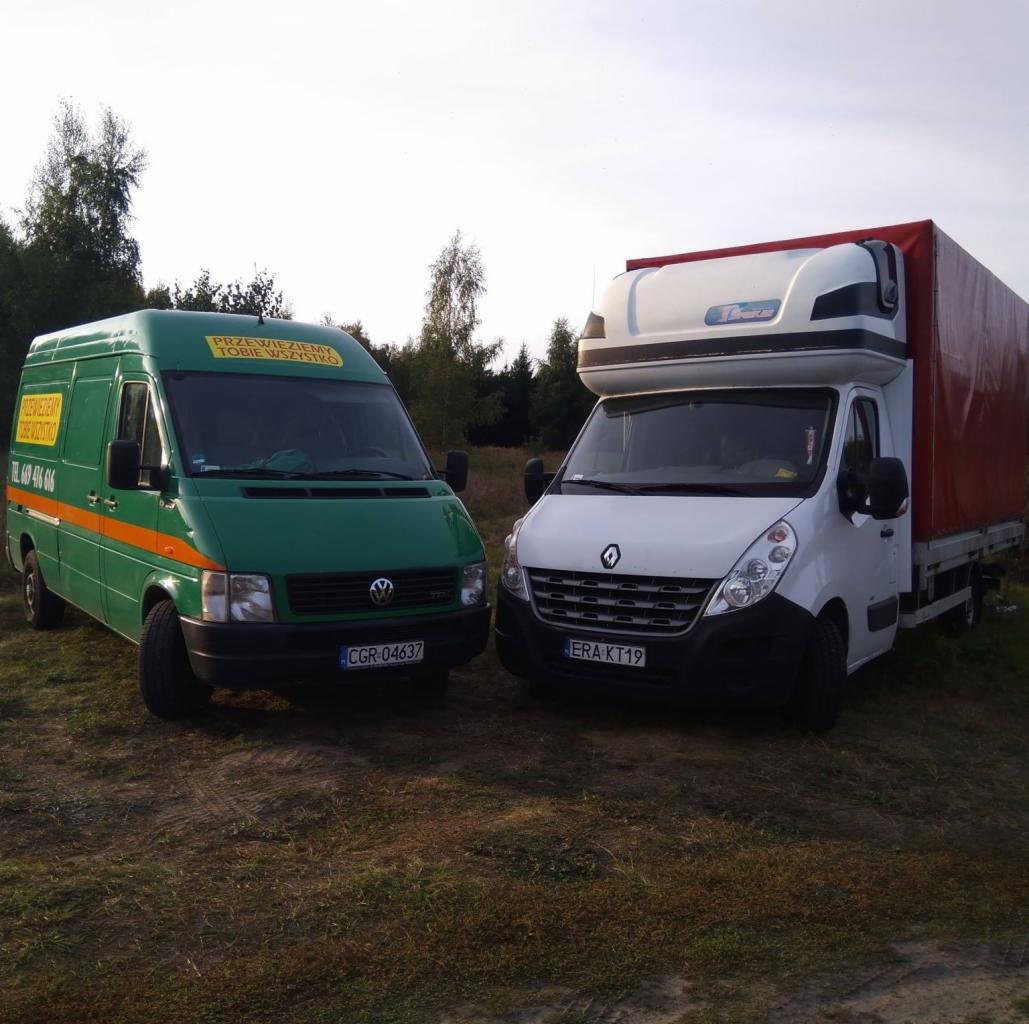 Dwa busy: zielony VW LT z pomarańczowym pasem i napisem reklamowym oraz biały Renault Master z plandeką, stojące na trawiastym terenie pod lasem.