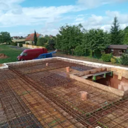 Zbrojenie stropu domu jednorodzinnego z widocznymi elementami szalunku i pionowymi rurami wentylacyjnymi, w tle dwa samochody dostawcze i zabudowa jednorodzinna.