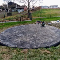 Okrągły plac z szarej kostki brukowej ułożony na trawniku, w tle domy jednorodzinne i sprzęt budowlany, zagęszczarka wibracyjna na kostce.