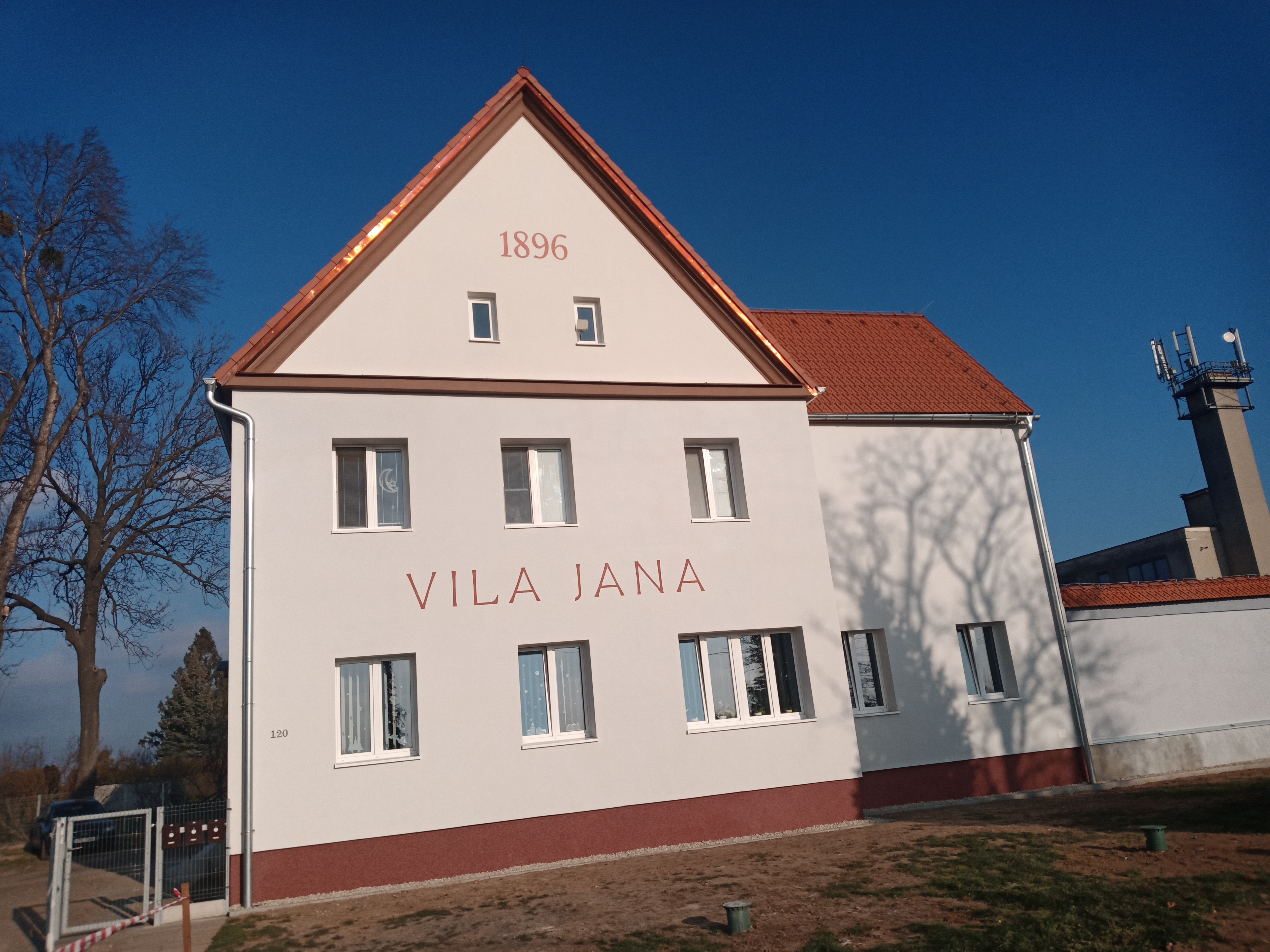 Odnowiony, dwukondygnacyjny budynek z jasną elewacją, czerwonym dachem i napisem 'Vila Jana' oraz '1896', usytuowany na tle błękitnego nieba i drzew bez liści.