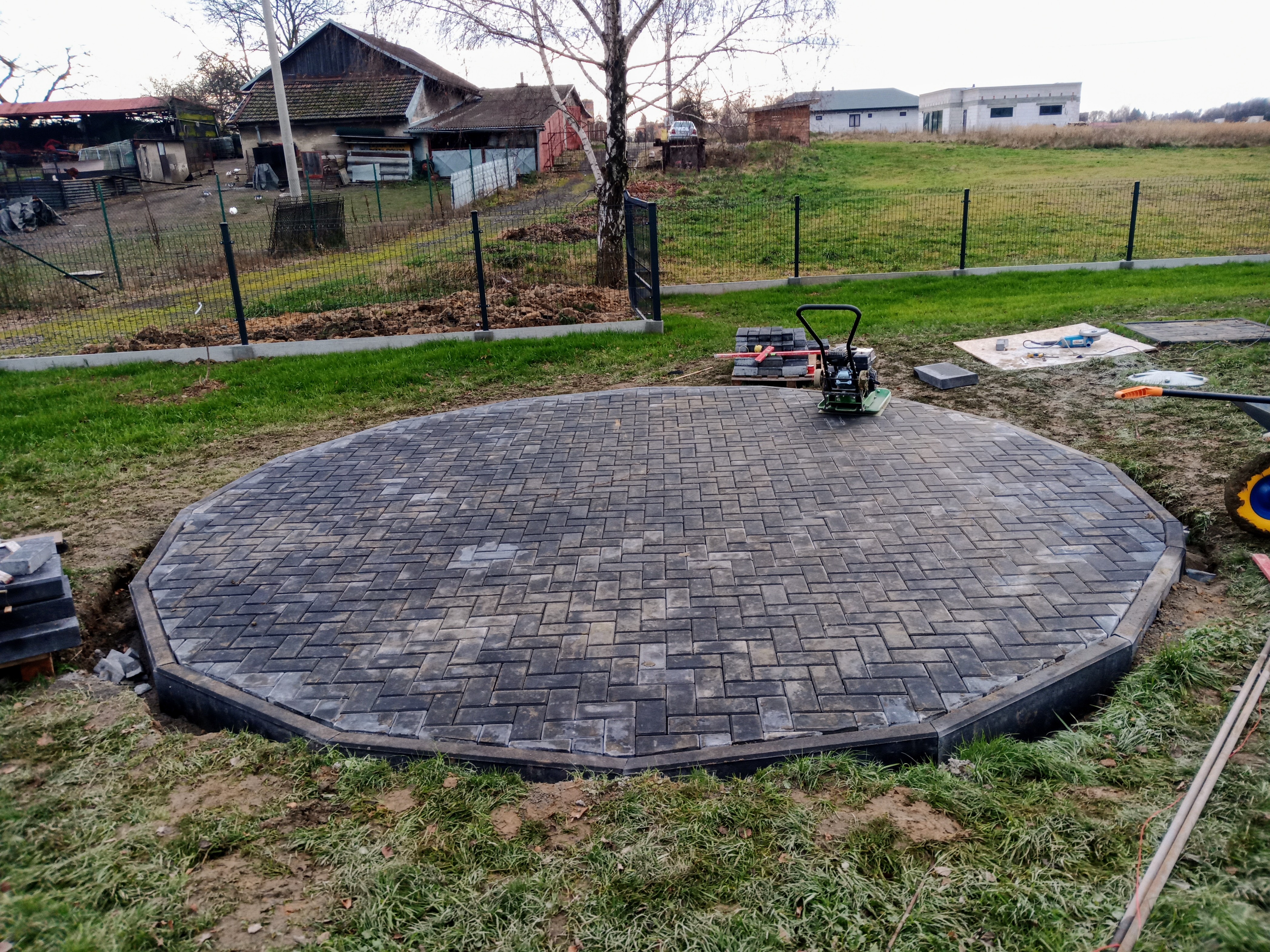 Okrągły plac z szarej kostki brukowej ułożony na trawniku, w tle domy jednorodzinne i sprzęt budowlany, zagęszczarka wibracyjna na kostce.