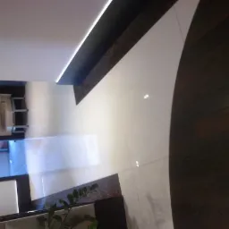 Widok z góry na fragment wnętrza z białą błyszczącą podłogą i ciemnym, drewnianym wykończeniem w łuk, z widocznym fragmentem rośliny doniczkowej w rogu.