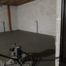 Wyrównywanie świeżo wylanej, ciemnoszarej posadzki w surowym pomieszczeniu z białymi ścianami z bloczków, widoczna zacieraczka mechaniczna.