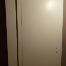 Białe, minimalistyczne skrzydło drzwiowe z widocznymi dwoma otworami na klamkę, zamontowane w ościeżnicy w jasnym wnętrzu.