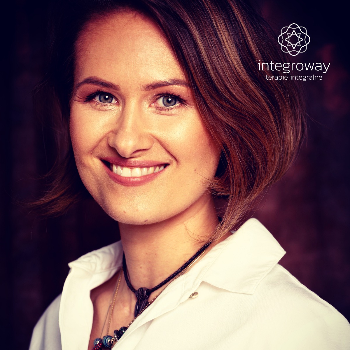 Portret uśmiechniętej kobiety z krótkimi brązowymi włosami, w białej koszuli i z naszyjnikami, na ciemnym tle, z widocznym logo 'integroway terapie integralne' w prawym górnym rogu.