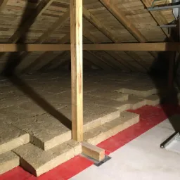 Poddasze z widoczną izolacją z wełny mineralnej ułożoną na czerwonej folii, drewniana konstrukcja dachu oraz komin ze stali nierdzewnej.