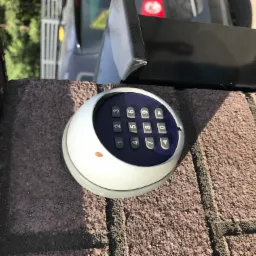 Zewnętrzny, okrągły panel kodowy domofonu z klawiaturą numeryczną, zamontowany na murze z cegły klinkierowej, widok z góry, obok czarnego pudełka z elektroniką domofonu.