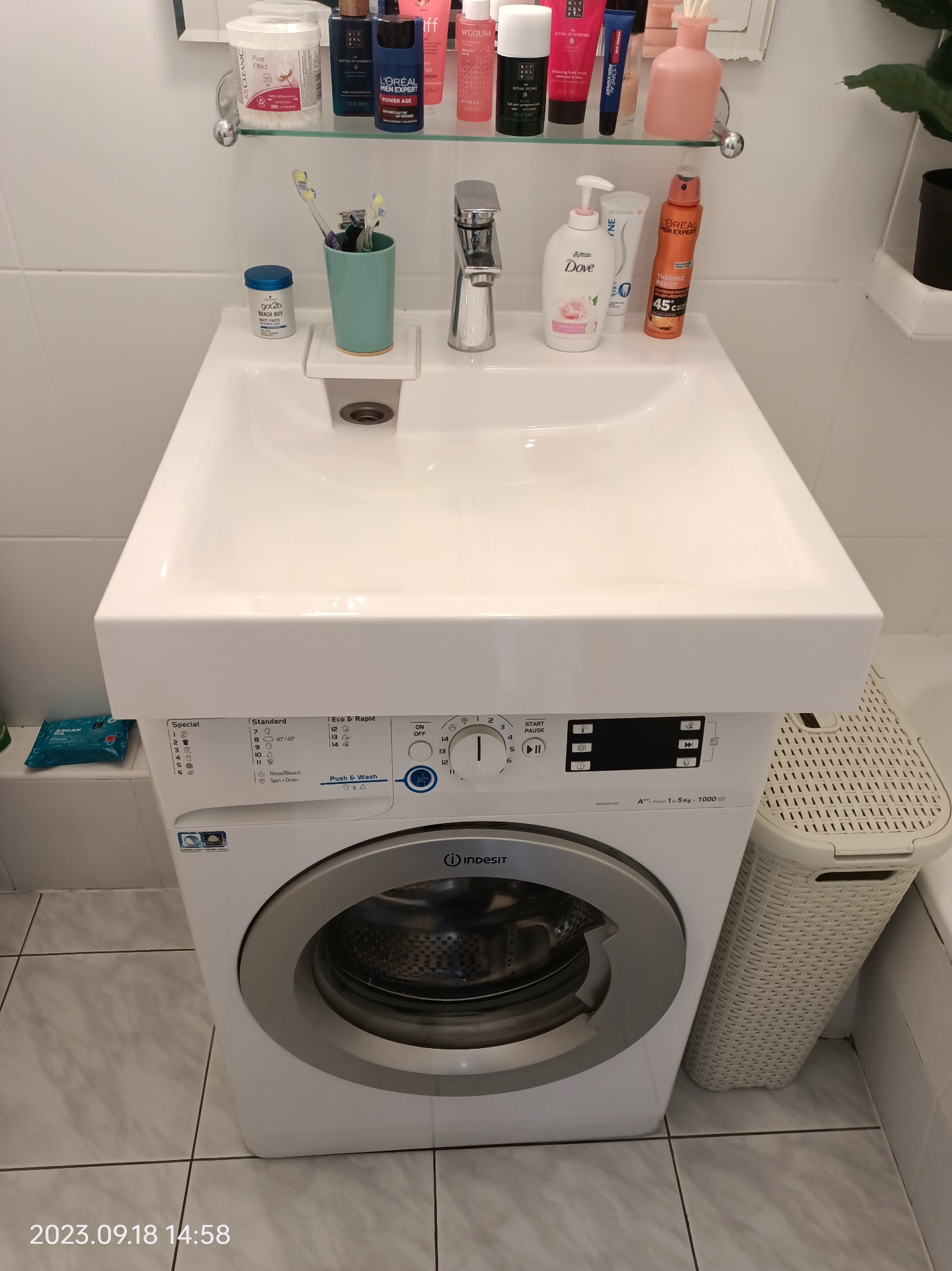 Umywalka nablatowa zamontowana nad pralką Indesit, z widocznymi kosmetykami i akcesoriami łazienkowymi na półce nad umywalką, oraz koszem na pranie obok pralki.