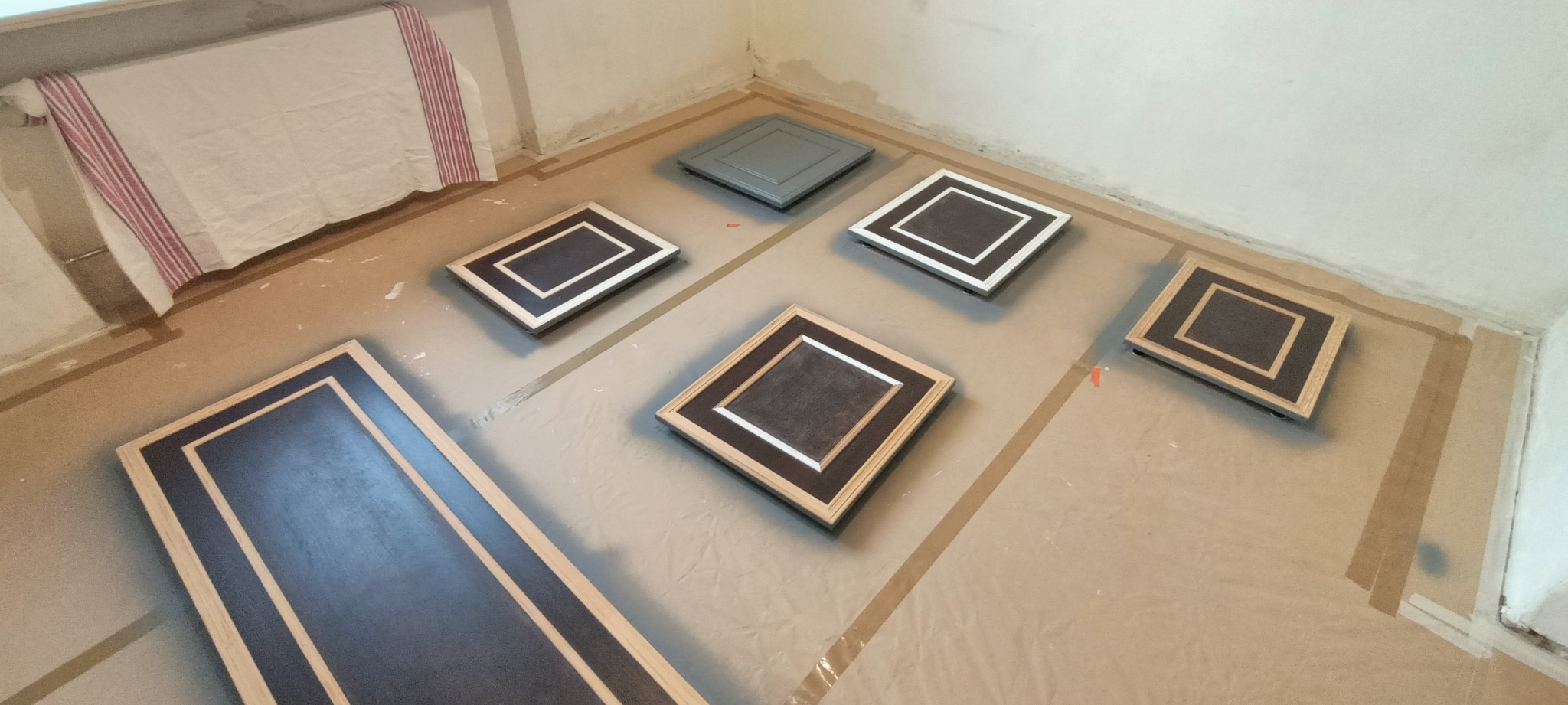 Fronty szafek przygotowane do natryskowego malowania, leżące na zabezpieczonej folią podłodze w rogu pomieszczenia.