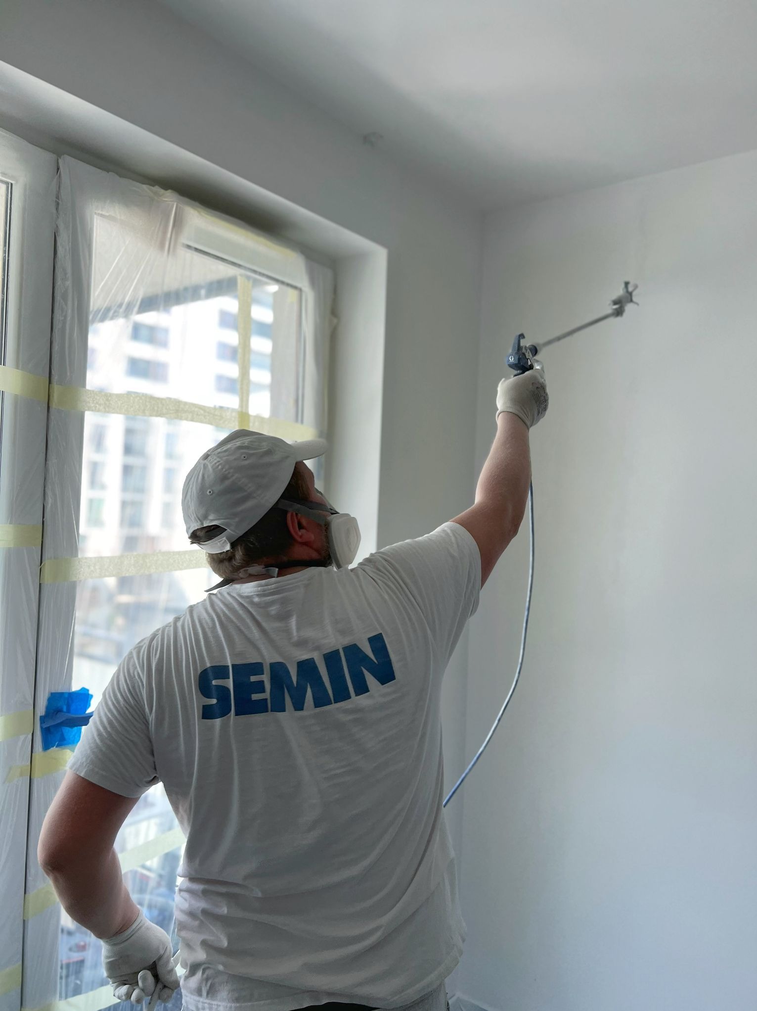 Pracownik w masce i czapce, w koszulce z napisem SEMIN, maluje ścianę natryskowo w jasnym pomieszczeniu. Okno zabezpieczone folią i taśmą malarską.