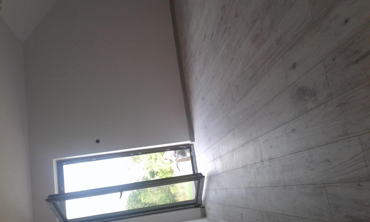 Nowoczesne okno dachowe uchylone, widok na zieleń z zewnątrz, jasna drewniana podłoga i białe ściany w wykończonym wnętrzu.