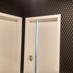 Glazurtech - Dwa białe, częściowo zamontowane drzwi z ościeżnicami, zabezpieczone niebieską folią, na tle ściany pokrytej tapetą w geometryczny wzór kostek.