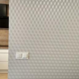 Glazurtech - Jasna ściana z geometrycznym wzorem 3D w odcieniach szarości, obok fragment białej kuchni z czarnym okapem i blatem z naturalnego drewna.