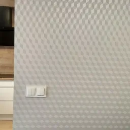 Jasna ściana z geometrycznym wzorem 3D w odcieniach szarości, obok fragment białej kuchni z czarnym okapem i blatem z naturalnego drewna.