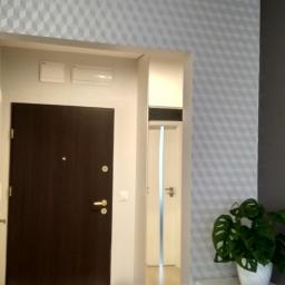 Glazurtech - Nowoczesne wnętrze mieszkania po remoncie: ciemnobrązowe drzwi wejściowe, ściana w geometryczne wzory 3D, widoczny fragment korytarza z białą szafką i drzwiami, dekoracyjna roślina Monstera...