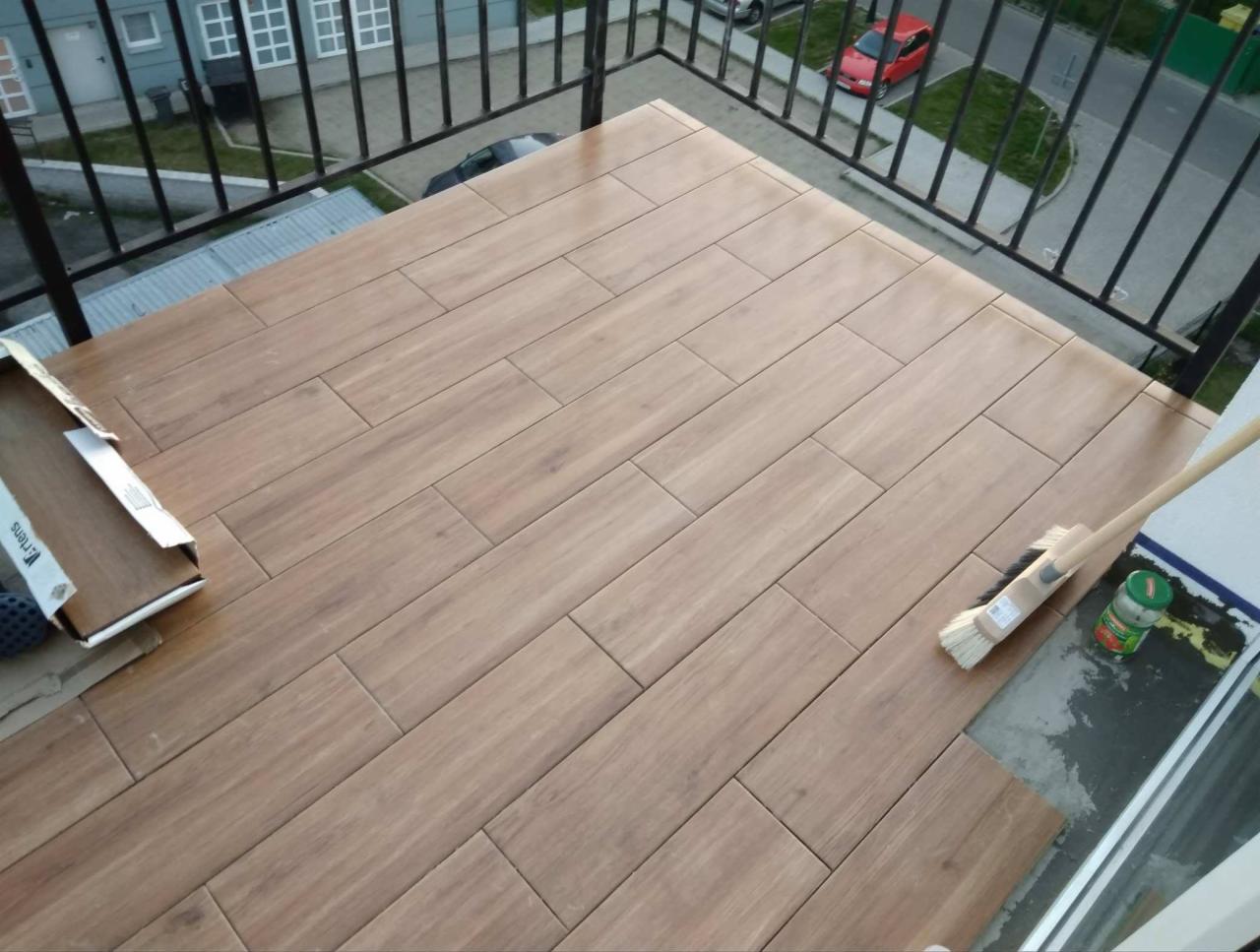 Układanie drewnopodobnych płytek na balkonie, widoczny karton z płytkami, szczotka i słoik na krawędzi balkonu, widok z góry na częściowo wykończoną podłogę.