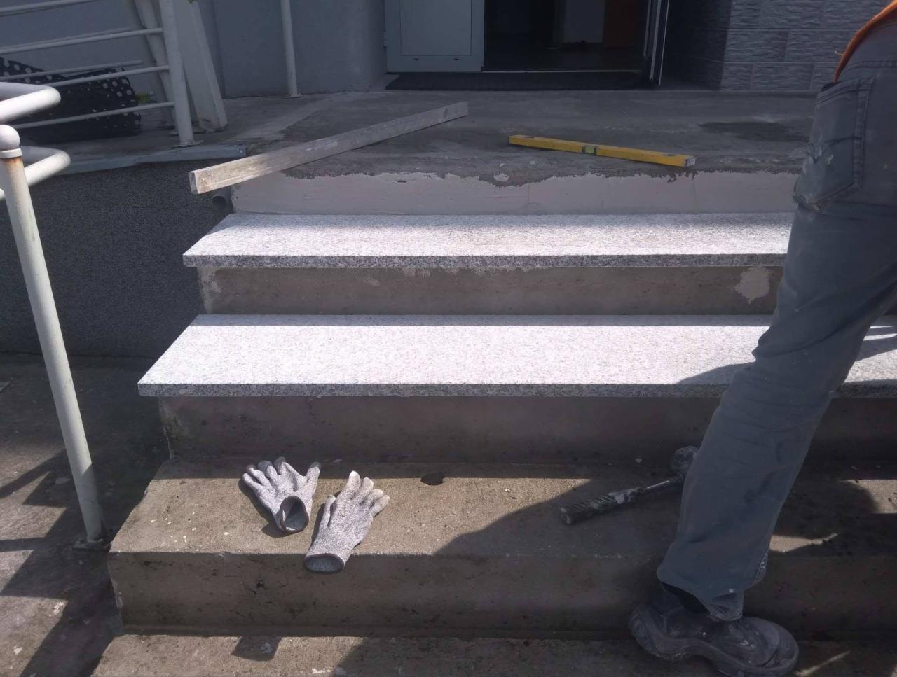 Montaż granitowych płyt na schodach wejściowych, widoczne narzędzia pracy: poziomica, młotek i rękawice robocze.
