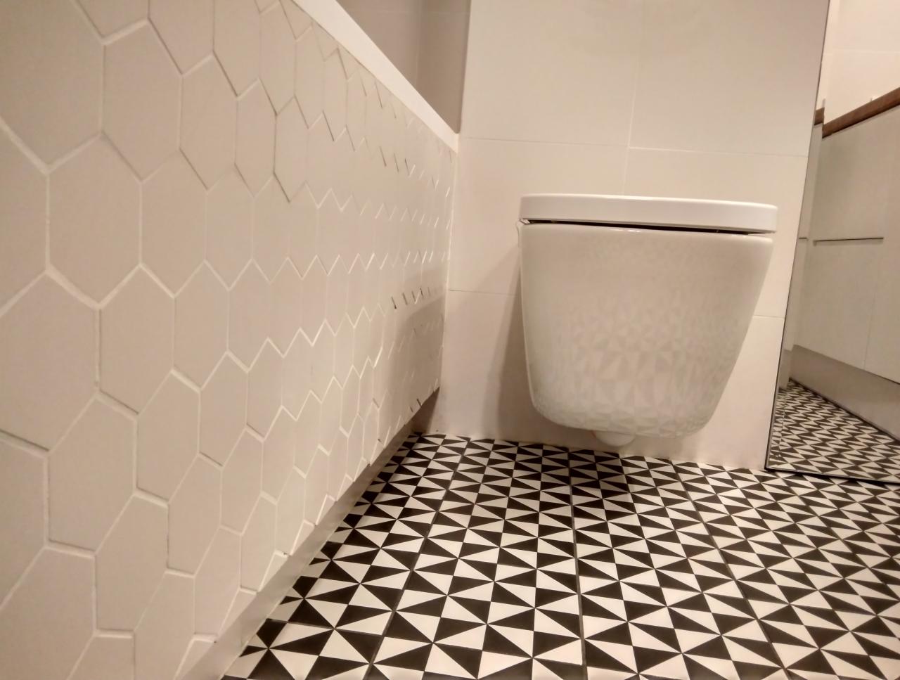 Nowoczesna łazienka z wiszącą toaletą, heksagonalnymi płytkami na ścianie i geometryczną posadzką w czarno-białe trójkąty.