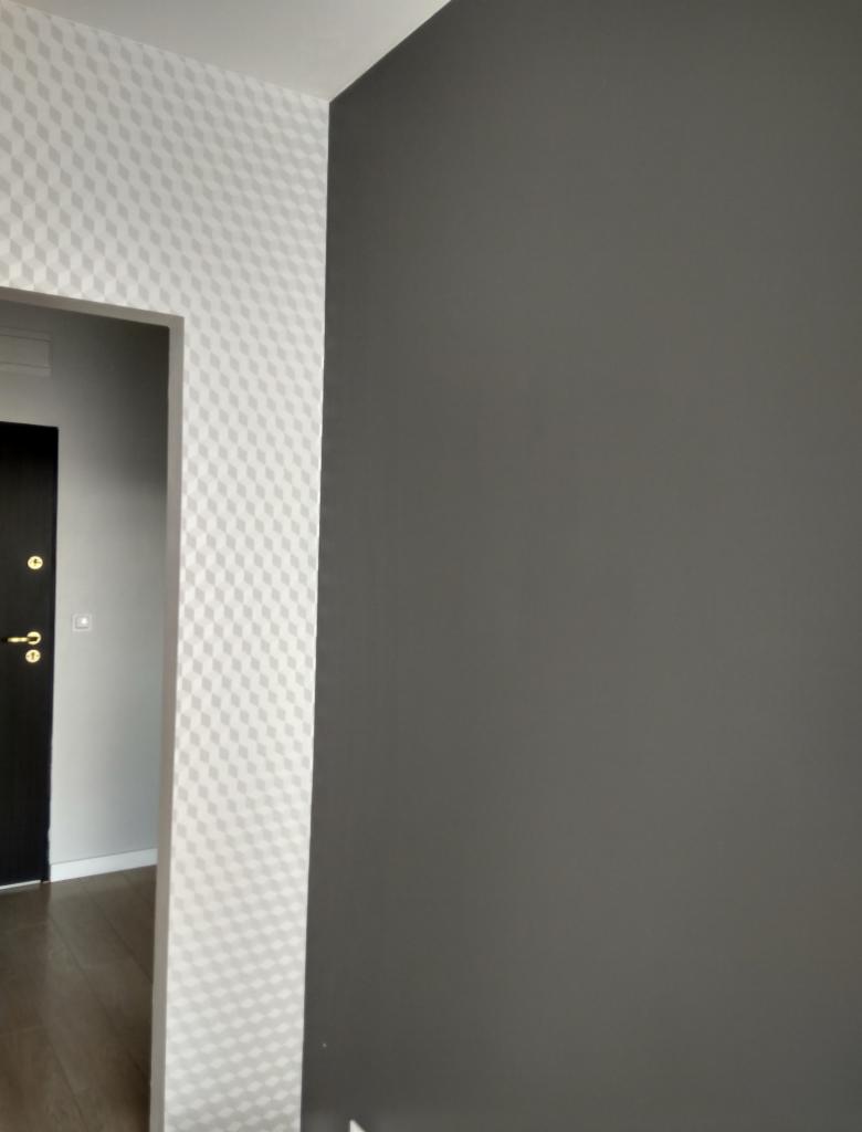 Fragment korytarza z pomalowaną na szaro ścianą i ścianą przy wejściu do pokoju z białą tapetą w geometryczny wzór rombów.