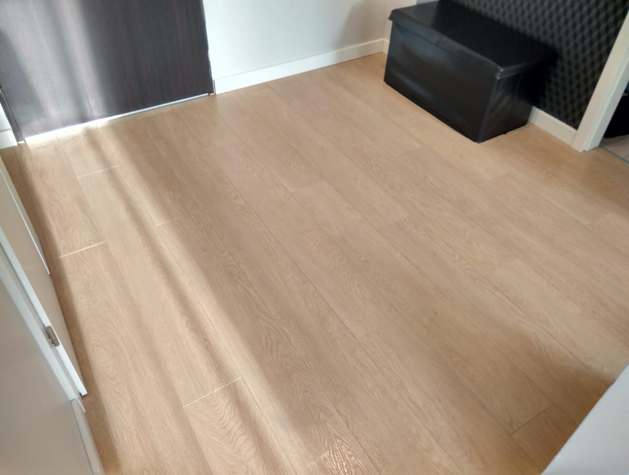 Jasne panele podłogowe ułożone wzdłuż, widoczny fragment drzwi, czarna pufa ze schowkiem i panel akustyczny na ścianie.