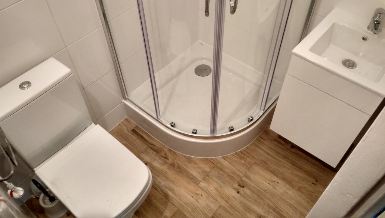 Ukończona łazienka z kabiną prysznicową narożną, toaletą i umywalką z szafką, wykończona w białych płytkach i drewnopodobnych panelach podłogowych.