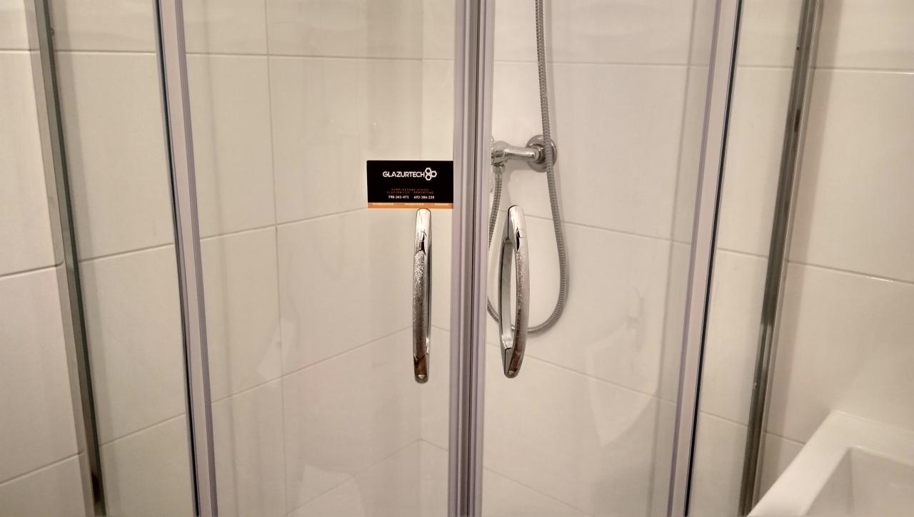 Szklana kabina prysznicowa z chromowanymi uchwytami i słuchawką prysznicową na tle białych płytek; wizytówka firmy 'Glazurtech' przyklejona do szyby.