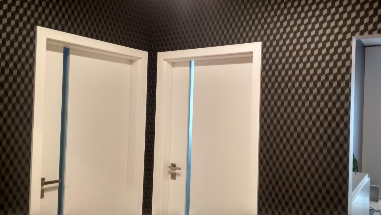 Dwa białe, częściowo zamontowane drzwi z ościeżnicami, zabezpieczone niebieską folią, na tle ściany pokrytej tapetą w geometryczny wzór kostek.