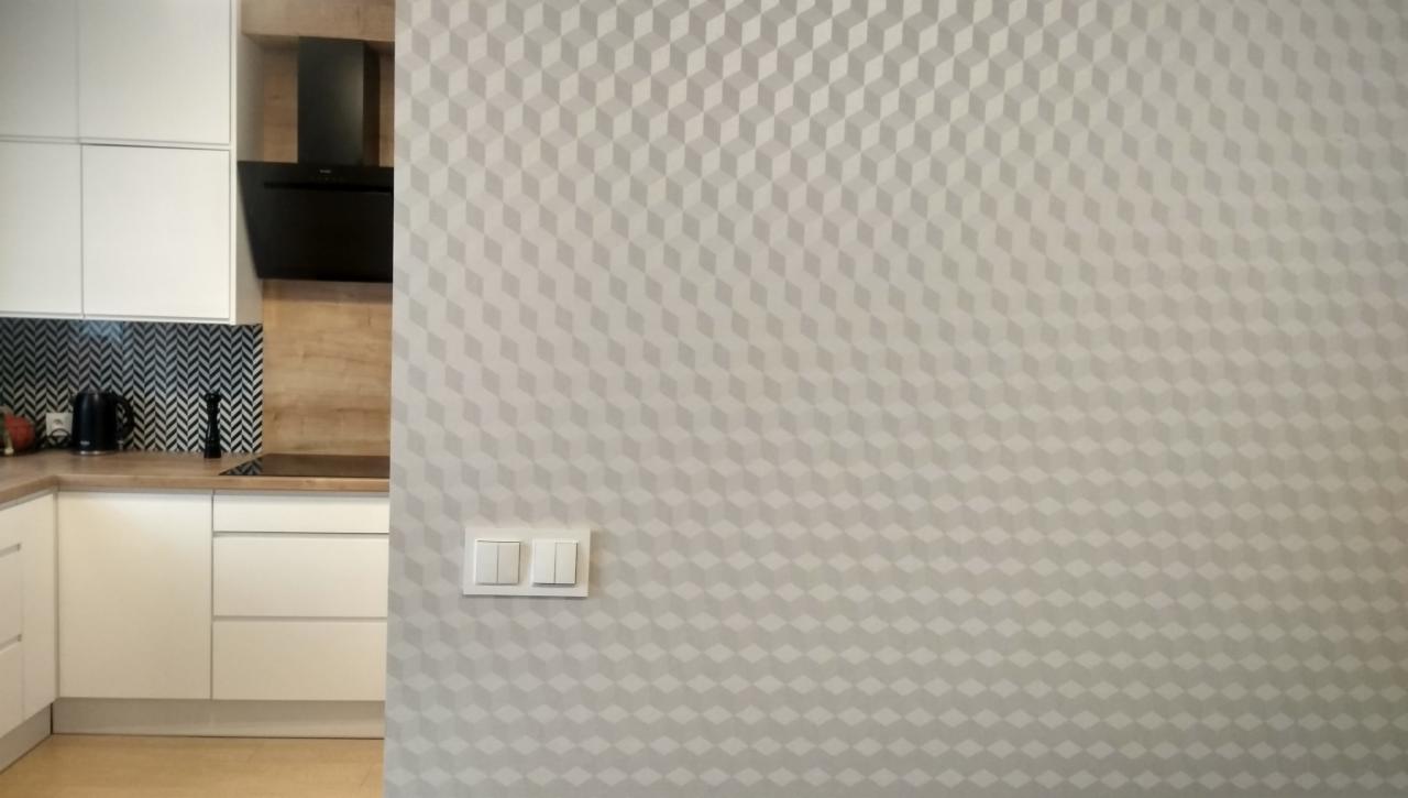Jasna ściana z geometrycznym wzorem 3D w odcieniach szarości, obok fragment białej kuchni z czarnym okapem i blatem z naturalnego drewna.