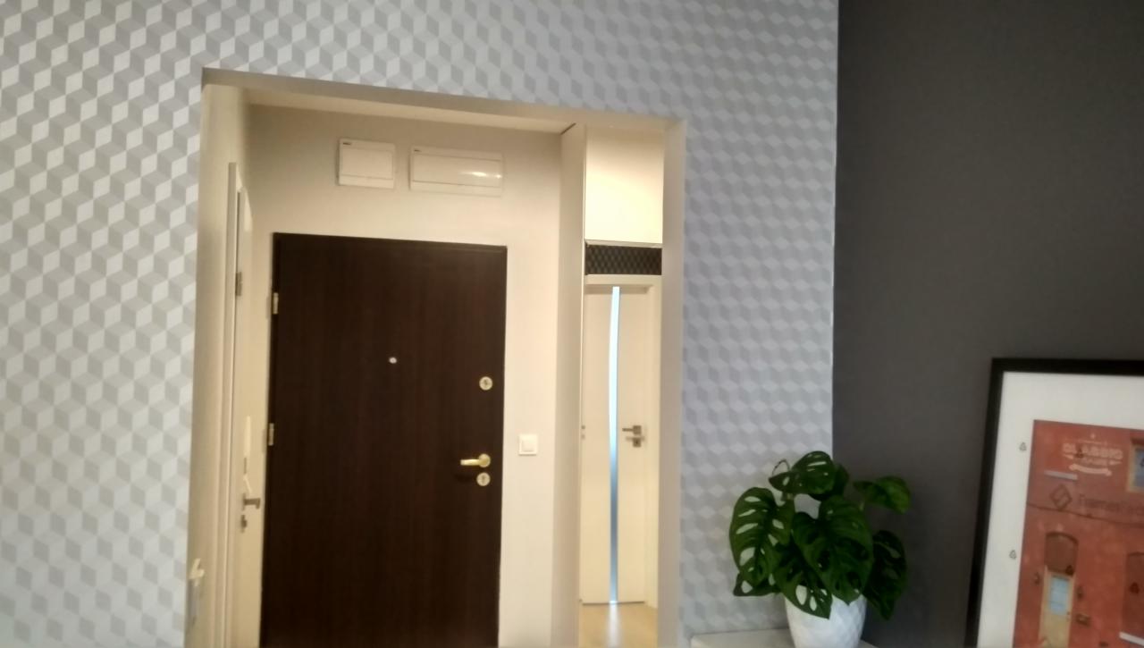 Nowoczesne wnętrze mieszkania po remoncie: ciemnobrązowe drzwi wejściowe, ściana w geometryczne wzory 3D, widoczny fragment korytarza z białą szafką i drzwiami, dekoracyjna roślina Monstera...