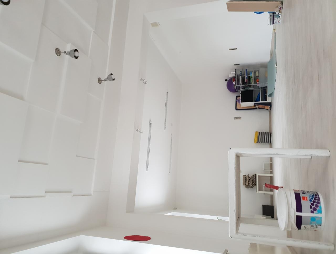 Białe, odświeżone wnętrze z widoczną strukturą ściany, oświetleniem punktowym i akcesoriami malarskimi, w tym wiadro z farbą i stolik malarski.