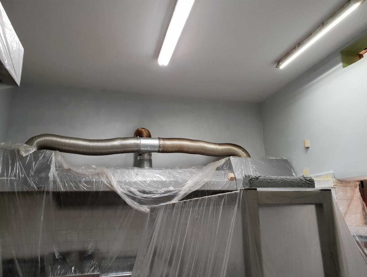 Metalowy przewód wentylacyjny przymocowany do sufitu, zabezpieczone folią meble i wałek malarski, świeżo pomalowana ściana w odcieniach szarości.