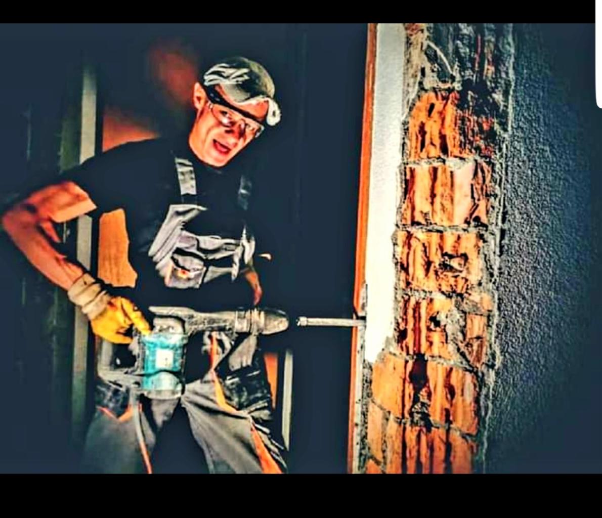 Mężczyzna w okularach i kombinezonie roboczym wierci w ścianie z cegły za pomocą wiertarki udarowej, oświetlony lampą czołową.