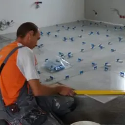 Mężczyzna w pomarańczowej koszulce i szarym kombinezonie poziomuje nową szarą płytkę podłogową za pomocą żółtej poziomicy, widoczne niebieskie klipsy dystansowe, żółty odkurzacz Stanley w rogu...