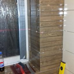 Piotr Stachowiak - Układanie płytek imitujących drewno na ścianie w łazience, widoczne krzyżyki dystansowe, poziomica, butelka napoju i narzędzia glazurnicze na parapecie okna zabezpieczonego folią.