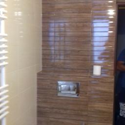 Piotr Stachowiak - Nowoczesna łazienka z wiszącą toaletą, brązowe płytki imitujące drewno na ścianie za toaletą, obok beżowe płytki, biały grzejnik drabinkowy po lewej stronie.