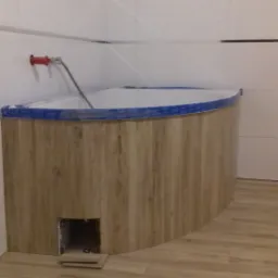 Obudowana wanna z drewnopodobnymi panelami, widoczny otwór rewizyjny i niezakończone układanie płytek podłogowych.