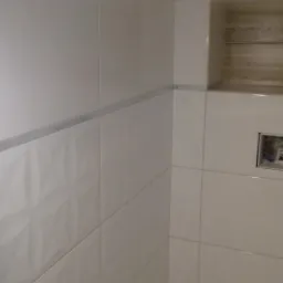 Ściana łazienki wyłożona białymi, błyszczącymi płytkami z dekoracyjnym pasem i wnęką z drewnopodobnym wykończeniem, widoczne elementy instalacji hydraulicznej.