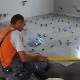 Piotr Stachowiak - Mężczyzna w roboczym kombinezonie i pomarańczowej koszulce układa szare płytki na podłodze, używając poziomicy i plastikowych krzyżyków dystansowych; w tle widoczne instalacje elektryczne...