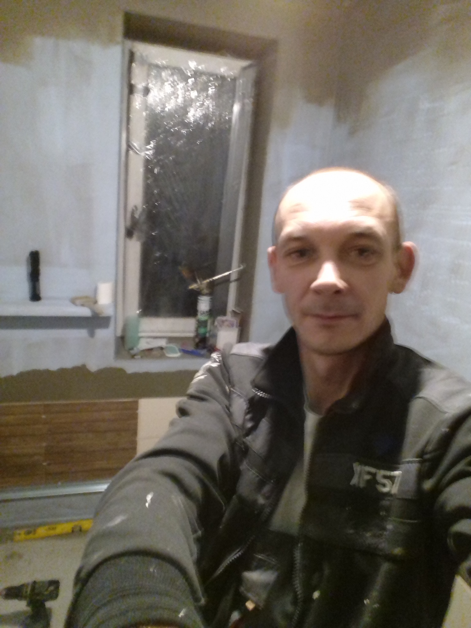 Autoportret mężczyzny w kurtce roboczej na tle ściany w trakcie układania brązowych płytek w łazience z oknem, widoczne narzędzia i materiały budowlane.