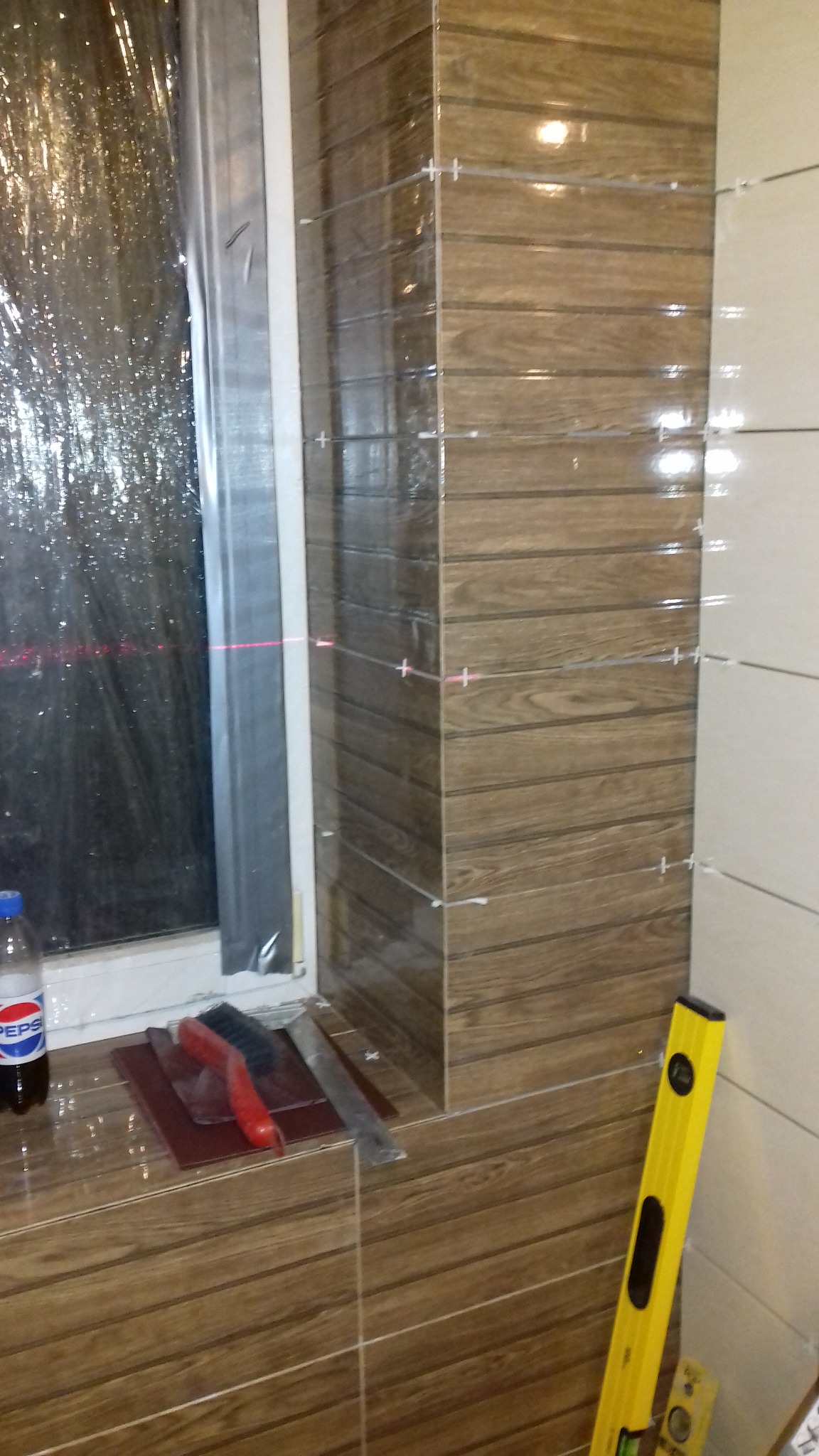 Układanie płytek imitujących drewno na ścianie w łazience, widoczne krzyżyki dystansowe, poziomica, butelka napoju i narzędzia glazurnicze na parapecie okna zabezpieczonego folią.