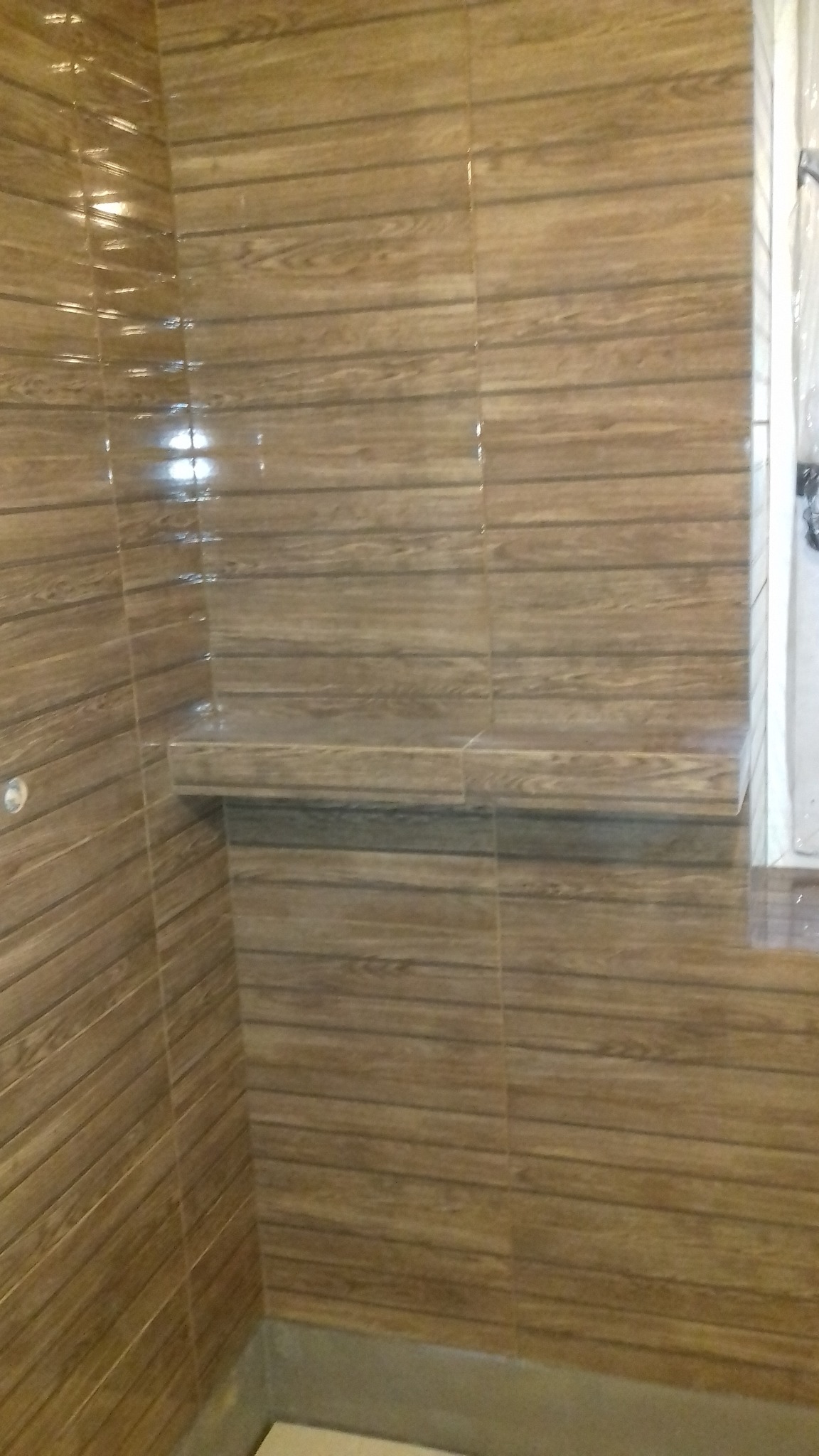 Wykończona łazienka z brązowymi płytkami imitującymi drewno, z półką w rogu kabiny prysznicowej, widoczne delikatne refleksy światła na powierzchni płytek.