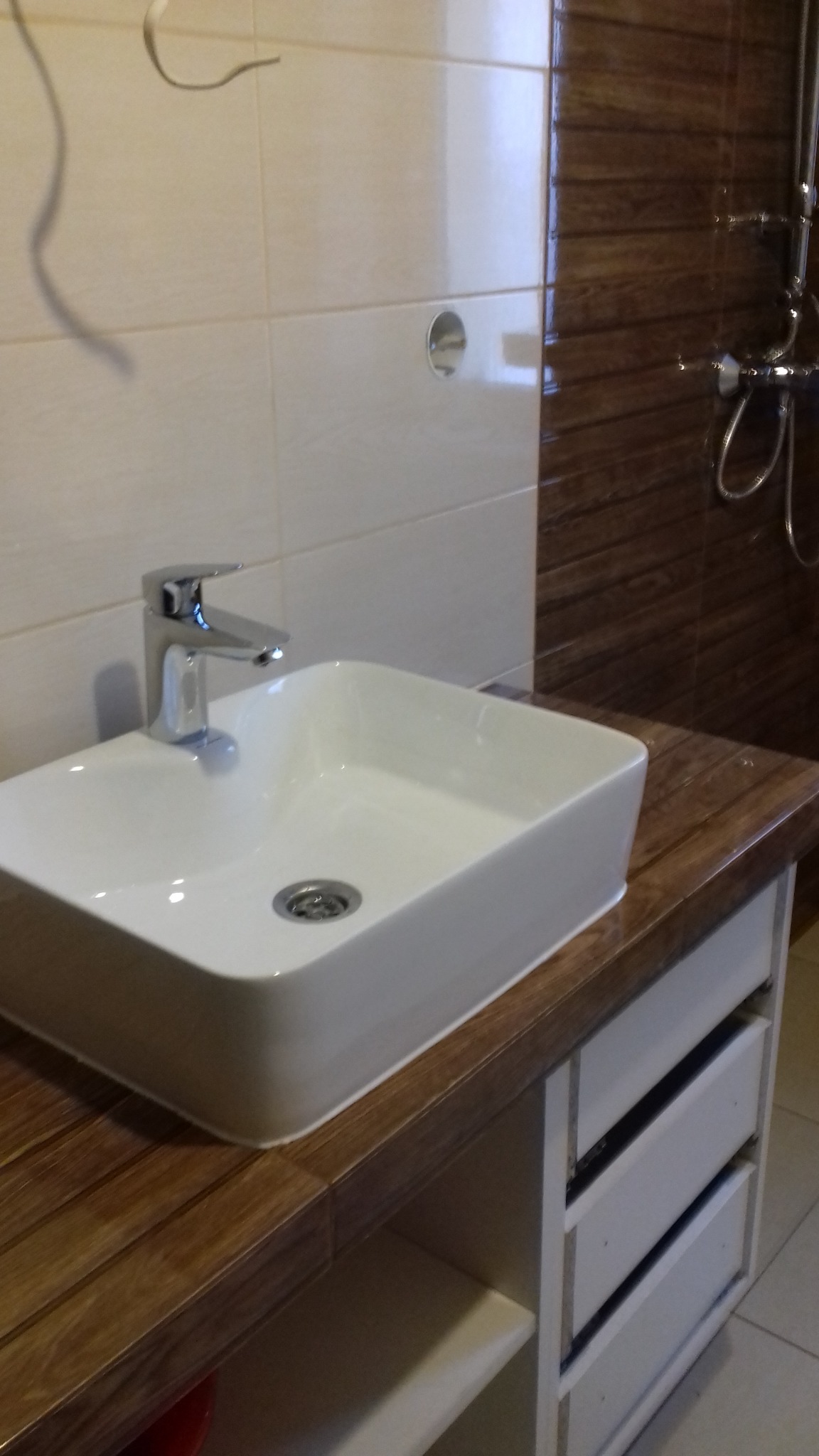 Biała, prostokątna umywalka nablatowa z chromowaną baterią, osadzona na drewnopodobnym blacie szafki z białymi szufladami w łazience z beżowymi i brązowymi płytkami.